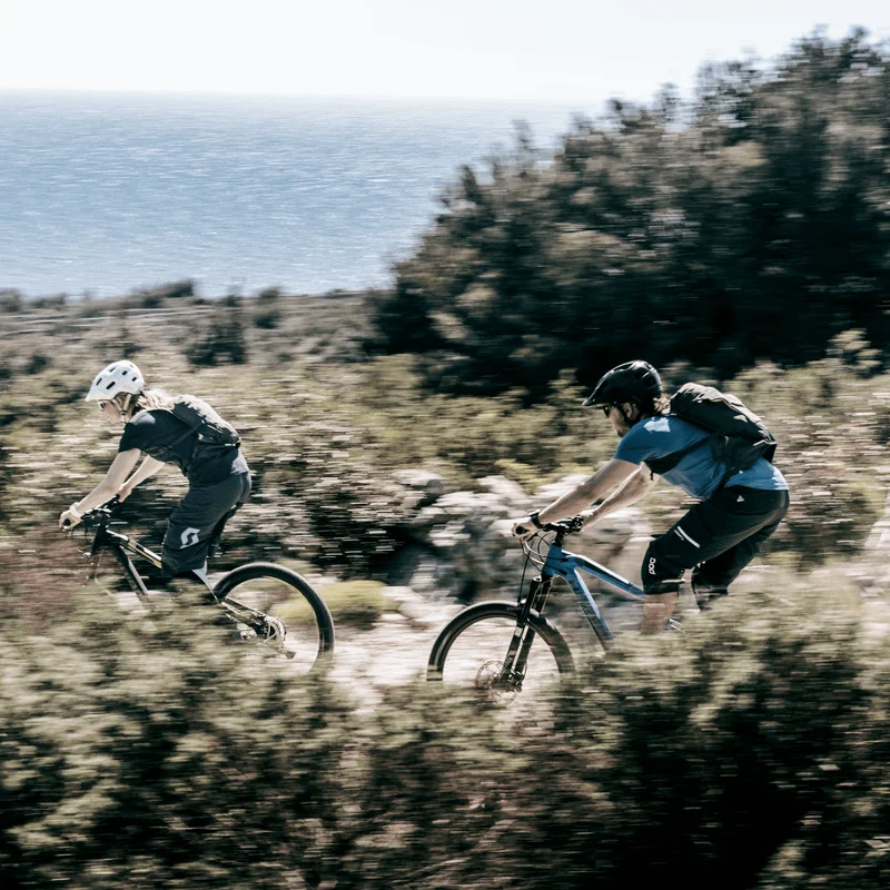 By the ocean, two people go mountain biking down a mtb trail with a Thule Vital hydration pack.