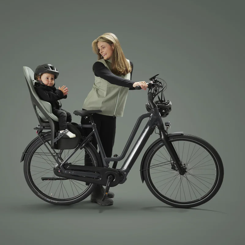 Eine Frau steht bei ihrem Fahrrad und schaut zurück zu ihrem Kind, das in einem Kindersitz auf dem Fahrrad sitzt.