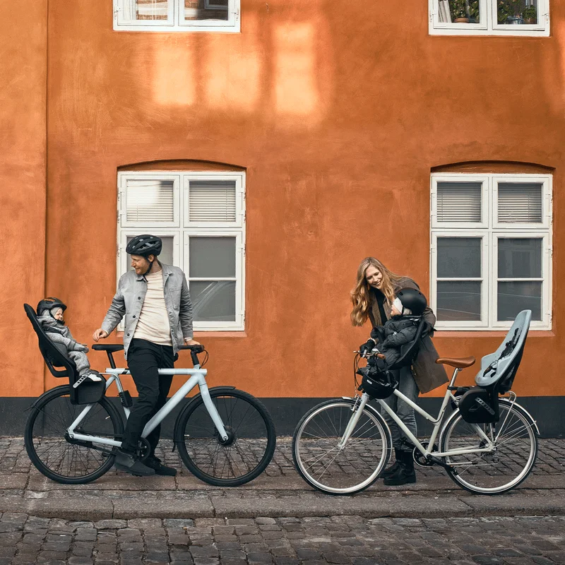 Neben einer orangefarbenen Wand stehen zwei Eltern mit Fahrrädern und Kindern in Thule Yepp 2 Maxi und Mini Kindersitzen.