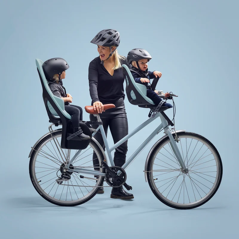 Eine Frau steht mit einem blauen Fahrrad, blauem Hintergrund und Kindern in einem blauen Thule Yepp 2 Maxi und Mini Fahrradkindersitz.