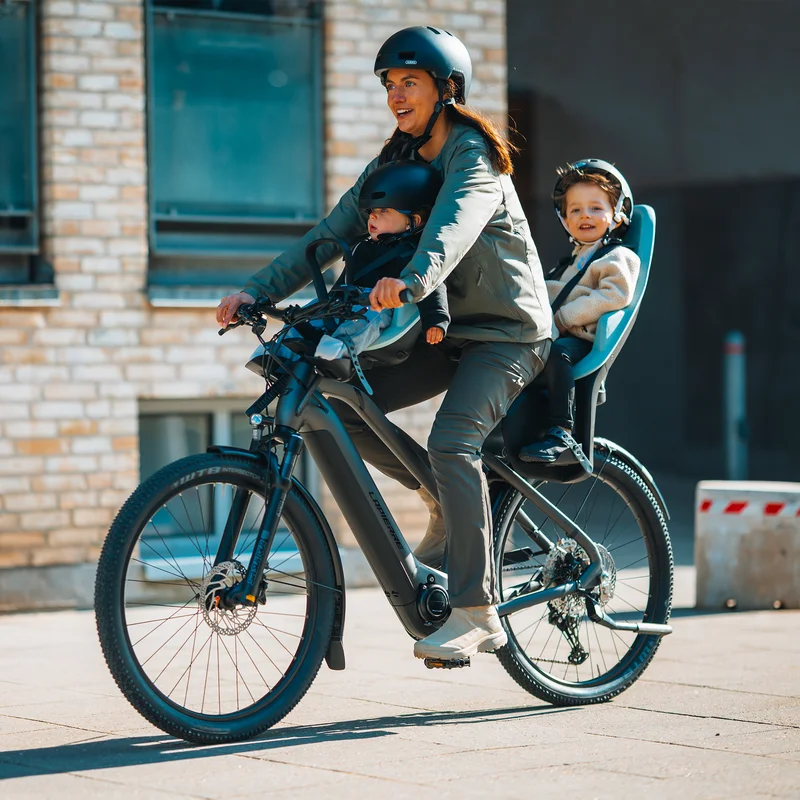 Stadtfamilie fährt mit zwei Kindern Fahrrad, die sicher sitzen, eins in einem vorderen und eins in einem hinteren Thule Yepp 2 Fahrradsitz.