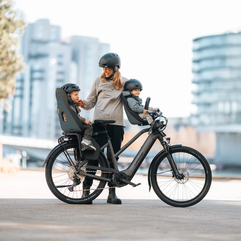 Erwachsener steht neben einem Fahrrad, das mit zwei Thule Yepp Nexxt 2 Fahrradsitzen für Kinder ausgestattet ist, einem vorne und einem hinten, während er mit zwei kleinen Kindern mit Helmen in einer städtischen Umgebung kommuniziert.