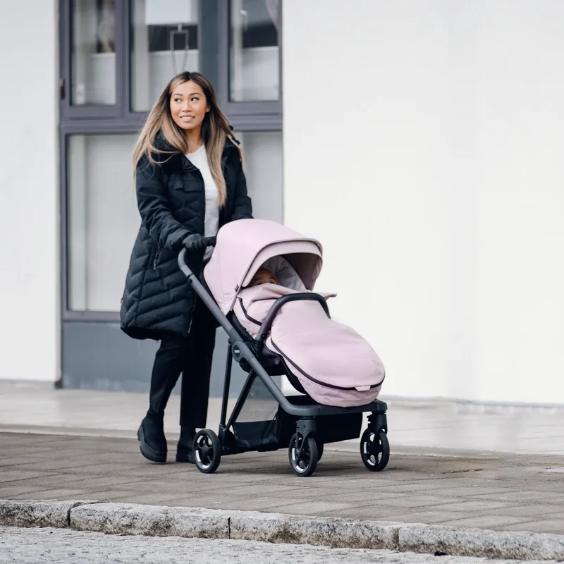 A woman in a jacket walks with her stroller and her baby in a pink Thule Stroller Footmuff.