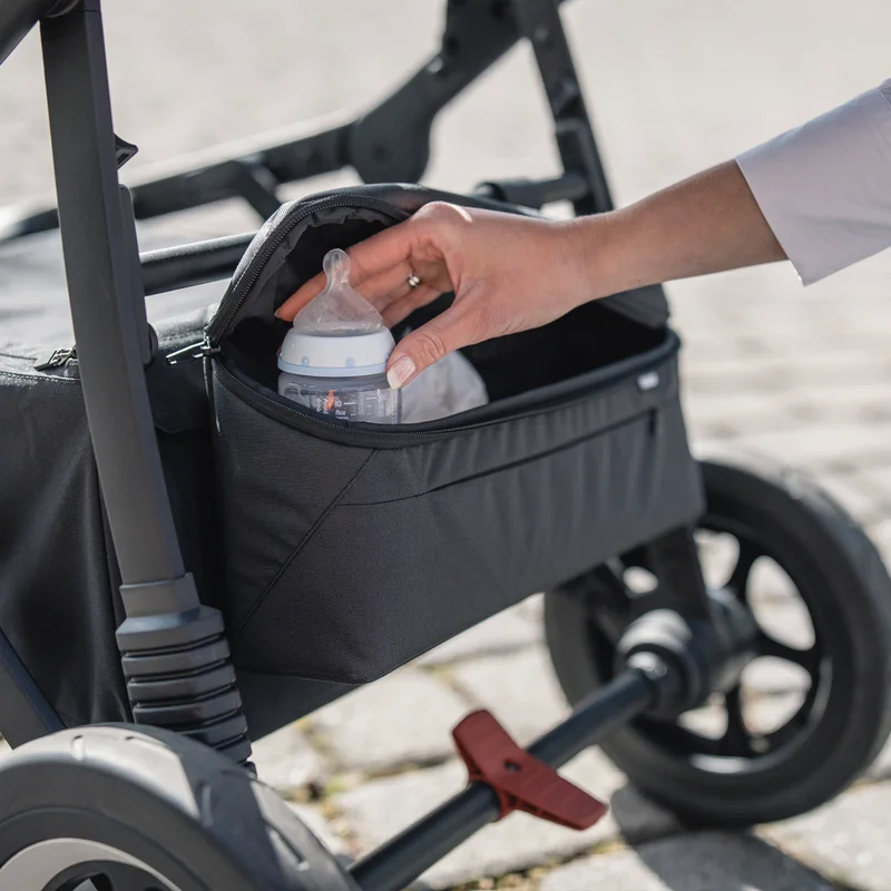 A hand places a bottle inside the Thule Stroller Organizer attached to the bottom of the stroller.